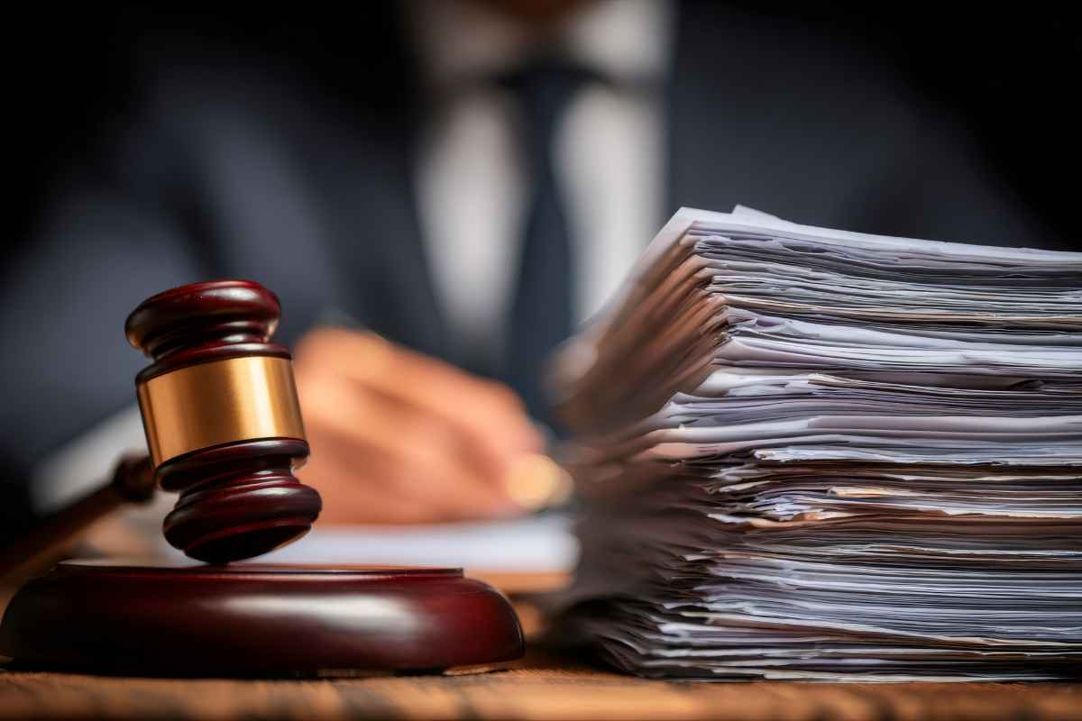 Martelo de juiz sobre base de madeira ao lado de uma pilha alta de documentos jurídicos, com figura desfocada de advogado ao fundo.