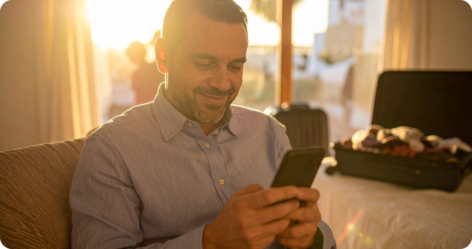 Homem sorrindo, usando smartphone no sofá com mala aberta ao fundo. Luz do sol.